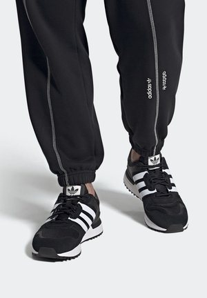 Stopy w czarno-białych butach sportowych Adidas z trzema paskami, połączone z czarnymi spodenkami dresowymi z białymi przeszyciami i logo Adidas.