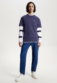 Lilla bomulls T-skjorte lagd over en hvit langermet skjorte med marineblå striper. Båret med blå jeans og hvite sneakers, stående på en ensfarget bakgrunn.