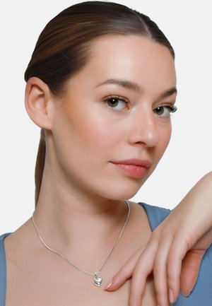 Giovane donna con capelli castani raccolti indossa una collana con ciondolo a forma di cuore in argento e una maglia blu, toccandosi delicatamente la spalla.