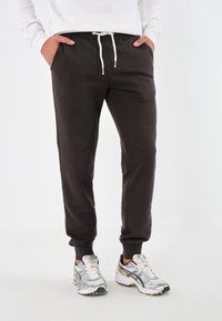 Dunkelbraune Sweatpants mit elastischen Bündchen und einem verstellbaren Kordelzugbund. Getragen mit silbernen und goldenen Sportschuhen.