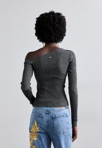 Γκρίζα ριγωτή μπλούζα με ανοιχτούς ώμους και μακριά μανίκια, με εφαρμοστή σχεδίαση και μικρή ετικέτα λογότυπου. Συνδυάζεται με μπλε τζιν που διαθέτουν λουλουδάτο σχέδιο.