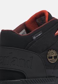 Czarne sneakersy z teksturowaną skórą zamszową, kontrastującym czerwonym wykończeniem i złotym akcentem z logo. Charakteryzują się grubą podeszwą i wyściełanym kołnierzem.