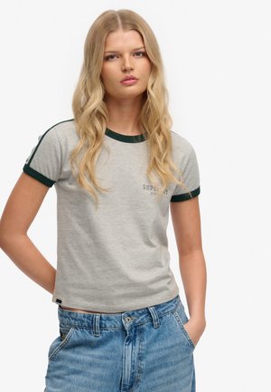 Ung kvinde med langt blondt hår står med den ene hånd i lommen, iført grå t-shirt med mørkegrøn kant og blå jeans.