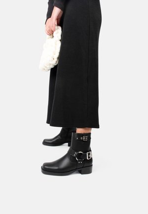 Person iført sorte læderstøvler til midt på læggen med sølvspænder, en lang sort nederdel og holder en chunky hvid strikket clutch taske.