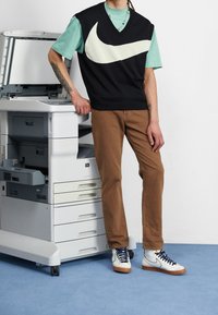Personne portant un gilet Nike noir, une chemise turquoise, un pantalon marron et des baskets Nike blanches se tenant à côté d'une imprimante de bureau ouverte sur un tapis bleu.