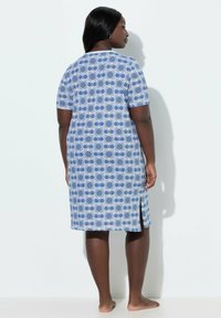 Mujer de piel oscura y cabello negro, vestida con un vestido de manga corta hasta la rodilla con patrones circulares azules y blancos, de pie descalza frente a una pared.