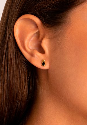 Orecchini d'oro con una pietra nera a forma di goccia, caratterizzati da un dettaglio decorativo a spirale, indossati nel lobo accanto a capelli castani.