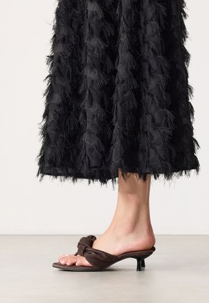 Blote voet met bruine suède kitten heel-sandalen met strikdetail, onder een zwarte gestructureerde midi-rok.