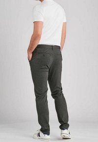 Hombre de pie con las manos en los bolsillos, vistiendo pantalones gris oscuro, camiseta blanca de manga corta y zapatillas blancas con rayas azul marino y rojas.