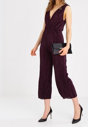 Femme portant une combinaison sans manches plissée de couleur violet profond avec des jambes larges raccourcies, tenant une pochette noire et portant des talons hauts noirs.