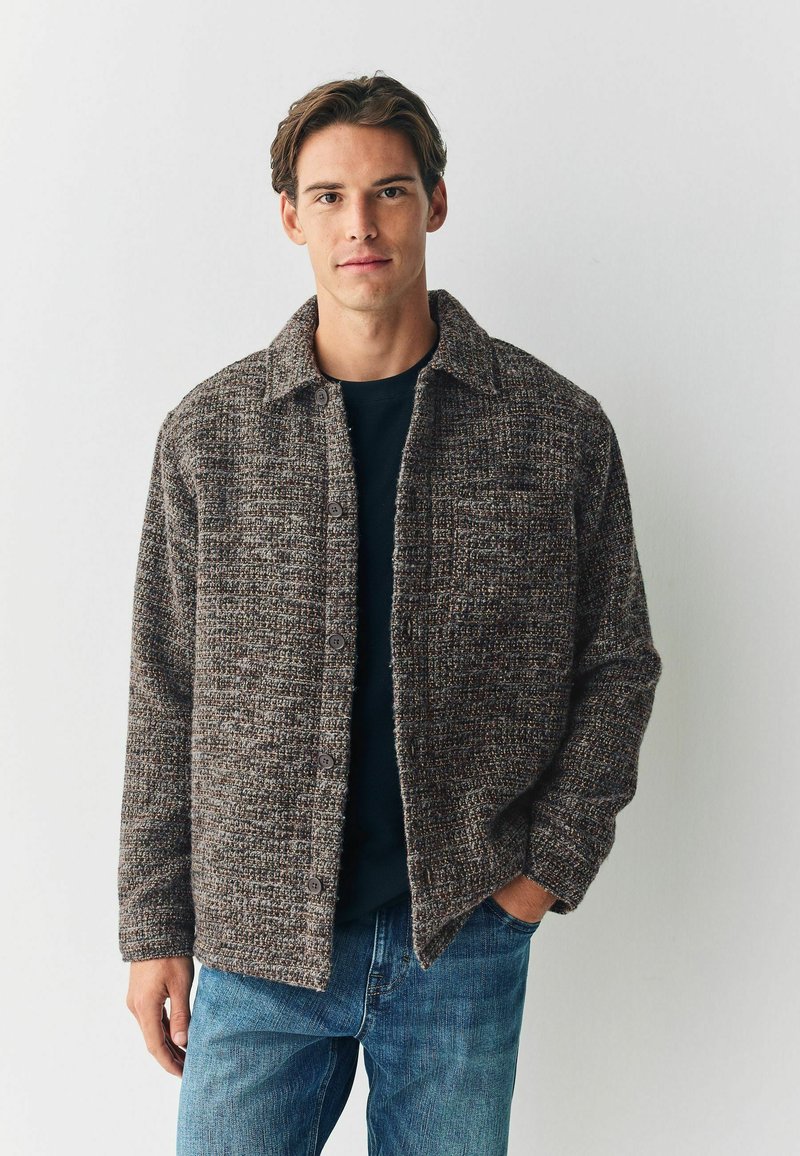 Chaqueta marrón texturizada con cuello, cierre de botones y dos bolsillos, superpuesta sobre una camiseta oscura y combinada con jeans azules.