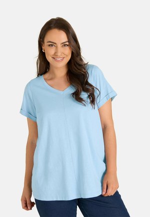 Woman with long dark hair smiling, wearing a light blue short-sleeve V-neck top and dark pants against a plain background.