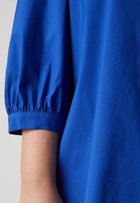 Blå bomullsblus med korta puffärmar, som har en mjuk textur och samlad elastik i manschetterna, vilket ger en lös passform.