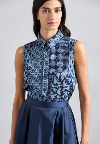 Blue denim sleeveless shirt with mixed patterns, featuring a button placket and contrasting sections; paired with a dark blue skirt.