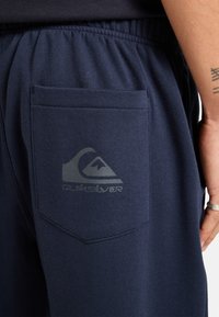 Marineblauwe broek van zachte stof, met een elastische tailleband, een achterzak en een subtiel grijs logo op de zak.