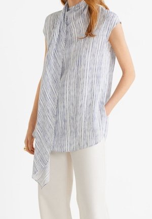 Femme portant un chemisier sans manches à rayures verticales bleues et blanches avec un drapé asymétrique à l'avant, associé à un pantalon blanc à jambes larges.