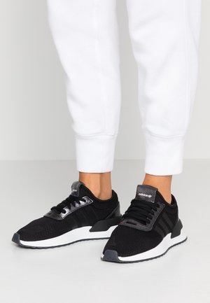Black Adidas sneakers with white soles worn with white pants on a person standing on a smooth light gray floor.