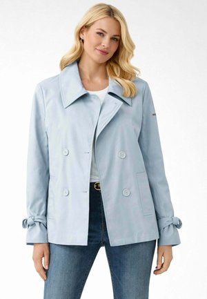 Donna che indossa un cappotto doppiopetto azzurro chiaro con ampio colletto e polsini annodati, abbinato a jeans blu e top bianco.