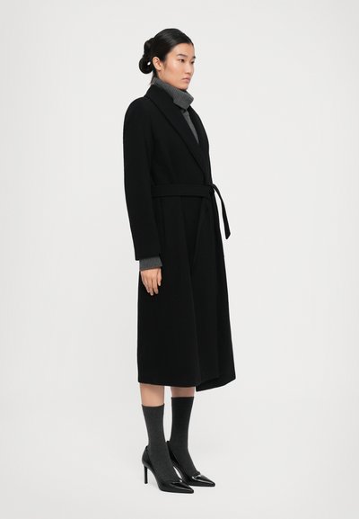 Woman wearing a long black belted coat, gray turtleneck sweater, gray socks, and black pointed high-heeled shoes standing sideways.