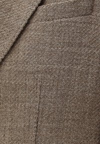 Strukturiertes braunes Gewebe zeigt ein Fischgrätmuster mit sichtbaren Nähten und einer Brusttasche. Das Material wirkt gewebt und langlebig.