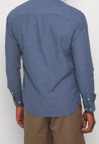 Mies, który ma na sobie niebieską koszulę z długim rękawem w białe kropki i brązowe spodnie, pokazany od tyłu na jednolitym tle.