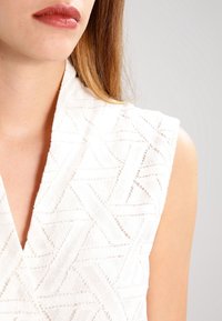 Nainen, jolla on vaaleanruskeat hiukset, pukee ylleen sleeveless-valkoisen tekstuuripaidan, jossa on geometrisia kuvioita. Hänen huulensa ovat hieman suljettuina ja tausta on neutraali.