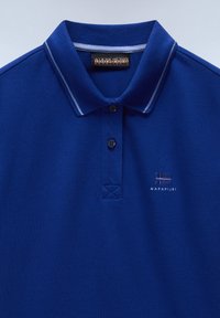 Blå pikétröja av texturerad tyg, med krage, tre knappar framtill och en liten broderad logotyp på bröstet.