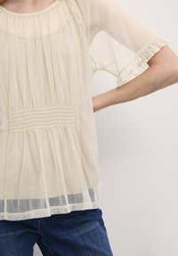 Gennemsigtig lys gul bluse med korte flæsekjoler, smock ved taljen og tekstureret prikket mønster hele vejen igennem. Matchet med blå jeans.
