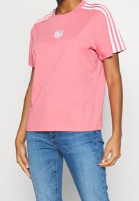 Rosa bomulls-t-shirt med korta ärmar, med tre vita ränder på axlarna och en broderad logotyp centrerad på bröstet.