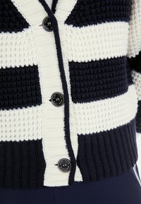 Gestrickte Strickjacke mit navyblauen und cremefarbenen Streifen, einem V-Ausschnitt, strukturiertem Stoff und einem schwarzen Knopf mit Logo-Detail an der Vorderseite.