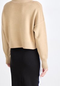 Beżowy, krótki sweter z prążkowanym dekolem i mankietami, o miękkiej fakturze i szerokich, opadających ramionach. Nosi się go na czarnej spódnicy.