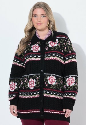 Kvinde iført en sort strikket cardigan med knapper med røde, hvide og grønne blomsterprint over en ternet skjorte.