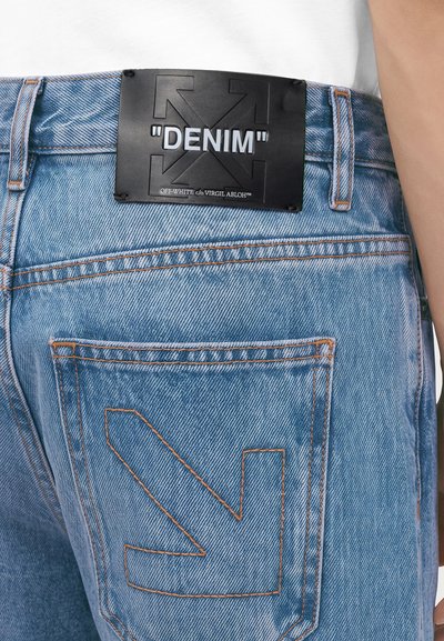 Jean en denim bleu clair avec coutures orange, patch en cuir noir portant l'inscription "DENIM" et un t-shirt blanc partiellement visible à la taille.