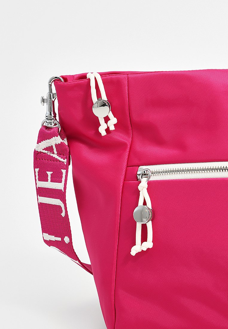 Rosa Nylon Tasche mit silberner Hardware, ausgestattet mit einer seitlichen Reißverschlusstasche und weißen Kordeln. Enthält einen gemusterten Riemen mit Text.