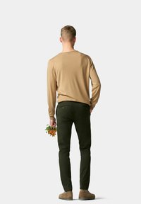 Beige overhemd met lange mouwen, donker groene slim-fit broek en bruine enkellaarzen. In de hand worden kleurrijke bloemen vastgehouden. Duidelijke achtergrond.