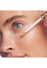 Zbliżenie twarzy kobiety, która nakłada płynny korektor za pomocą aplikatora w kształcie wędki; produkt ma jasny brzoskwiniowy odcień, skóra wygląda gładko i promiennie.