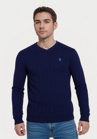Marineblauwe ribgebreide trui met V-hals, lange mouwen en een klein blauw logo op de borst. Gecombineerd met lichtblauwe spijkerbroek.