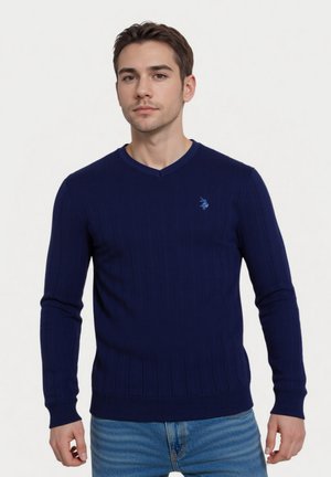 Jersey de punto acanalado en azul marino con escote en V, mangas largas y un pequeño logo azul en el pecho. Combinado con jeans de un azul claro.