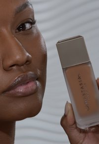 Bruine vloeibare foundation in een vierkante glazen fles, met een metalen beige dop. Etiket zegt "Anastasia Beverly Hills." Glad textuur zichtbaar.