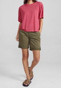 Červený top s krátkým rukávem a olivově zelené šortky s ohrnutými nohavicemi, doplněné hnědými sandály s dvojitými sponami a texturovaným materiálem.