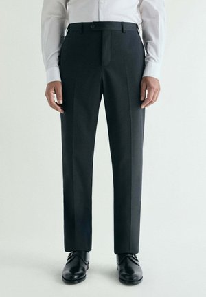 Hombre que lleva pantalones formales negros, zapatos de cuero negros y una camisa blanca de manga larga, de pie frente a un fondo claro y liso.