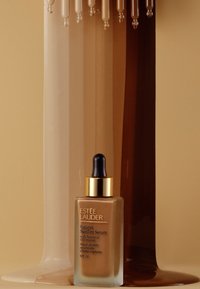 Sérum teint futuriste Estée Lauder dans un flacon en verre transparent avec un compte-gouttes, avec un capuchon doré et un sérum beige, posé sur un fond brun.