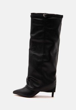 Botte haute en cuir noir avec un bout pointu, un talon moyen fin et une fine bride avec une petite boucle près du bord supérieur.