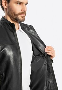 Zwarte leren jas met ritssluiting. Gladde textuur met een staande kraag en binnenzak, gedragen over een wit shirt.