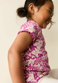 Rövid ujjú, lila virágmintás ing fodros szegéllyel és texturált szövettel, fehér virágmintákkal díszítve.