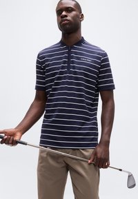 Άνδρας που φοράει μπλε ριγωτό πόλο πουκάμισο Calvin Klein και μπεζ παντελόνια κρατώντας ένα γκολφ ρόπαλο μπροστά σε απλό φόντο.