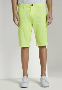 Limetkově zelené kraťasy vyrobené z lehkého materiálu, s rovnými nohavicemi, předními kapsami a černým knoflíkovým zapínáním. Nosené s bílými teniskami.