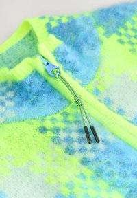 Detailný záber na chlpatý pletený kúsok oblečenia s modrým zipsom a neonovo zeleno-modrým károvaným vzorom, s čiernym koncovým ťahákom na šnúrku.