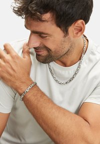 Zilveren metalen ketting en armband, met onderling verbonden schakels, gedragen om een blote arm en nek tegen een lichte achtergrond.