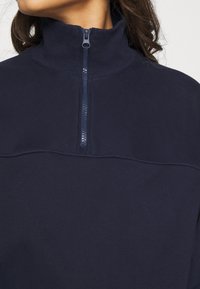 Pull bleu marine avec col montant et zip partiel, fabriqué en tissu doux. Présente une texture lisse et un design minimaliste.